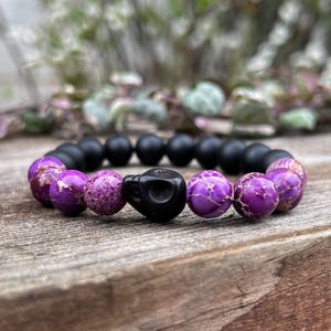 May include: A purple and black beaded bracelet with a skull charm. The bracelet is made with purple jasper and black onyx beads.