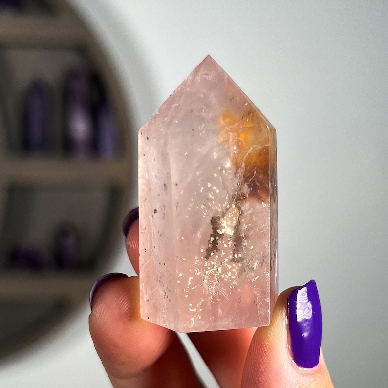 May include: A clear pink quartz crystal point with a golden inclusion. The crystal is faceted and has a pointed tip. It is a natural crystal and has a unique shape.