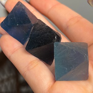 Puede incluir: Tres cristales de fluorita, uno negro y dos azul verdoso, en forma de octaedros. Los cristales est&aacute;n siendo sostenidos en una mano.