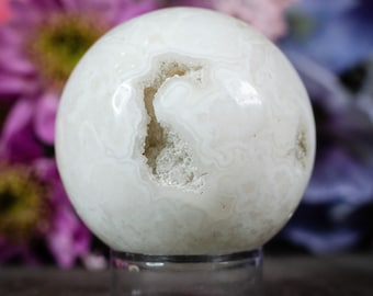 May include: White agate sphere with a natural druzy interior and a white, translucent exterior. The sphere is displayed on a clear stand.