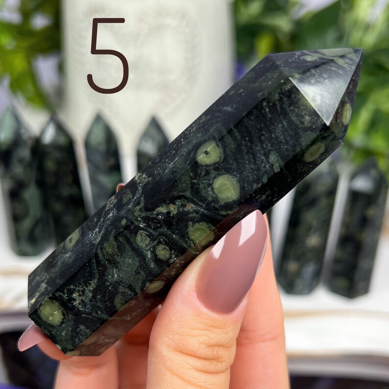 Pode incluir: Uma ponta de cristal verde escuro e preto com manchas brancas. O cristal tem a forma de uma pir&acirc;mide longa e fina. O cristal est&aacute; sendo segurado em uma m&atilde;o.