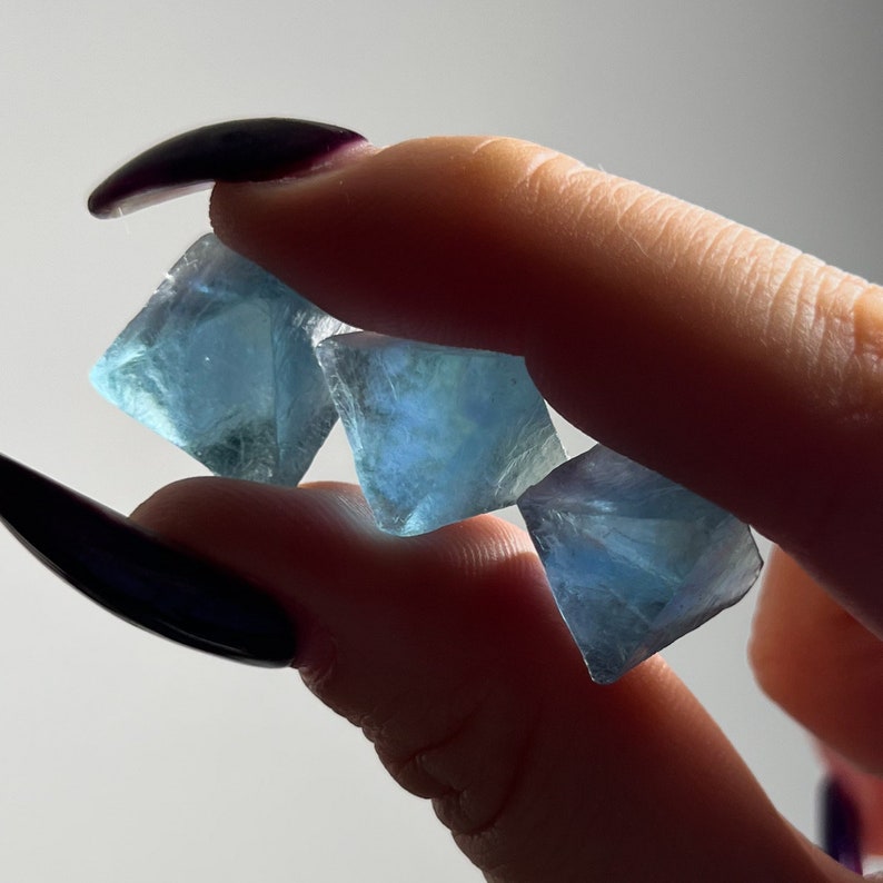 Puede incluir: Tres cristales de fluorita azules, cada uno con forma de pir&aacute;mide, se sostienen en una mano. Los cristales son transl&uacute;cidos y tienen un ligero brillo.
