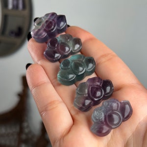 Könnte beinhalten: Fünf kleine, geschnitzte Lotusblüten-Anhänger aus violetter und grüner Fluorit. Die Anhänger sind in einer Reihe angeordnet, wobei der größte Anhänger oben und der kleinste Anhänger unten ist.