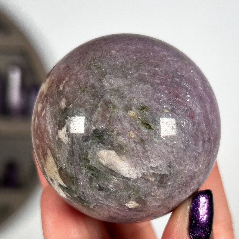 Puede incluir: Una gema esf&eacute;rica pulida, en tonos lavanda, gris y verde. La esfera tiene una superficie lisa y reflectante con vetas e inclusiones naturales. La bola de cristal se sostiene en una mano, mostrando su tama&ntilde;o y textura.