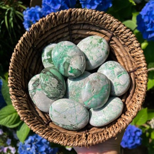 May include: A collection of green and white serpentine tumbled stones in a woven basket. The stones are smooth and polished, with a variety of patterns and textures.