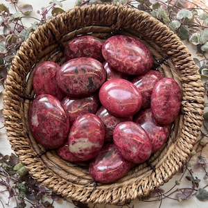 Peut inclure: Un panier en osier rempli de pierres polies de forme ovale. Les pierres sont d'un rouge profond avec des veines roses et brunes plus claires. Le panier est en matériau tissé brun clair.