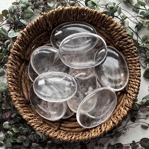 May include: A collection of clear quartz crystal palm stones in a woven basket. The stones are smooth and polished, with a translucent appearance.