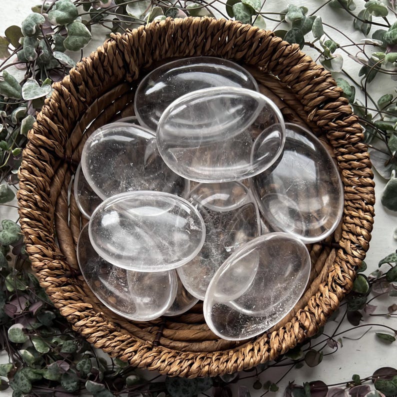 May include: A collection of clear quartz crystal palm stones in a woven basket. The stones are smooth and polished, with a translucent appearance.