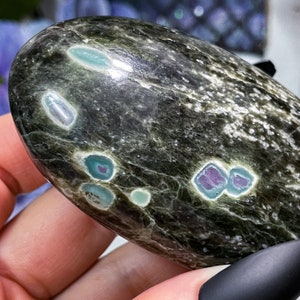 Może przedstawiać: Gładki, owalny kamień w kolorze zielonym z iryzującymi wtrąceniami. Kamień ma nakrapiany wzór i różne kolory, w tym zielony, niebieski, fioletowy i biały.