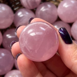 May include: A close-up of a pink rose quartz sphere held in a hand with dark purple nail polish. The sphere is smooth and translucent, with a soft pink hue. Other spheres are visible in the background.