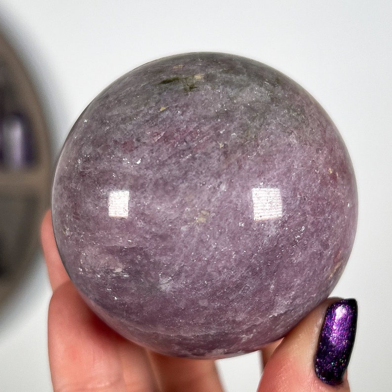 Puede incluir: Una piedra preciosa esf&eacute;rica pulida en tonos lavanda y gris. La bola de cristal tiene una superficie lisa con sutiles variaciones de color y textura. La esfera se sostiene en una mano, con un fondo blanco.