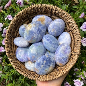 May include: A collection of blue celestite tumbled stones in a woven basket. The stones are smooth and have a light blue color with white veins.