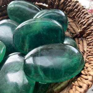 May include: A selection of polished, oval-shaped green fluorite stones. The stones are in a woven basket, displaying their translucent quality and varying shades of green. The smooth surfaces reflect light, highlighting the natural patterns within.