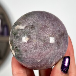 Puede incluir: Una gema esf&eacute;rica pulida, en tonos lavanda y gris, con sutiles inclusiones verdes y blancas. La superficie refleja la luz, mostrando su textura suave. Esta esfera decorativa es un art&iacute;culo de decoraci&oacute;n &uacute;nico.