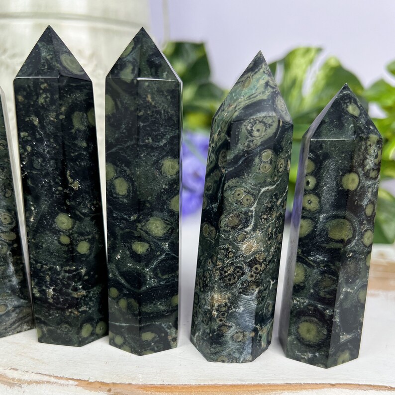 Pode incluir: Cinco pontas de pedra verde e preta salpicadas, tamb&eacute;m conhecidas como aventurina verde, est&atilde;o dispostas em uma fileira sobre uma superf&iacute;cie branca. As pontas s&atilde;o todas aproximadamente do mesmo tamanho e forma, e t&ecirc;m um acabamento liso e polido.