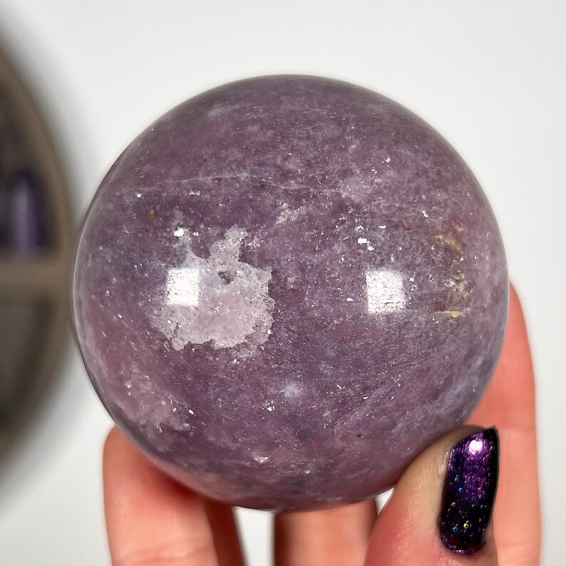 Puede incluir: Una gema esf&eacute;rica pulida, en tonos lavanda y p&uacute;rpura. La bola de cristal tiene una superficie lisa con sutiles variaciones de color y textura. La esfera se sostiene en una mano, mostrando su tama&ntilde;o y forma.