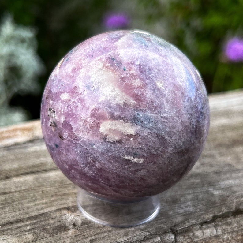Puede incluir: Una gema esf&eacute;rica pulida, en tonos lavanda, blanco y gris, que descansa sobre un soporte de acr&iacute;lico transparente. La superficie del orbe muestra un patr&oacute;n veteado, con sutiles variaciones de color y textura. El fondo est&aacute; borroso.