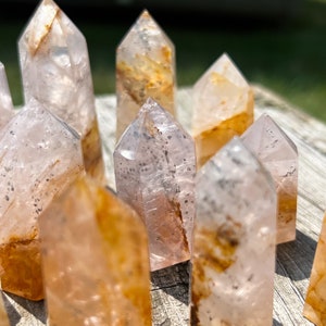 May include: A group of clear quartz crystal points with a light pink hue and brown inclusions. The points are arranged in a cluster on a wooden surface.