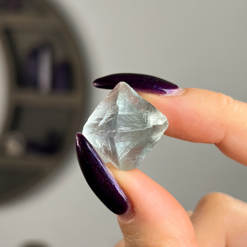 May include: A clear, faceted fluorite crystal with a slightly green hue. The crystal is shaped like a cube and has a smooth, polished surface.