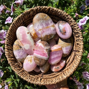 May include: A collection of pink and brown banded tumbled stones in a woven basket. The stones are smooth and polished, with a variety of patterns and textures.