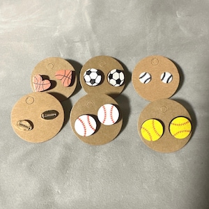 May include: Six pairs of wooden earrings with different sports ball designs. The earrings are displayed on small, round, brown paper cards. The designs include a basketball, a soccer ball, a baseball, a softball, and a football.