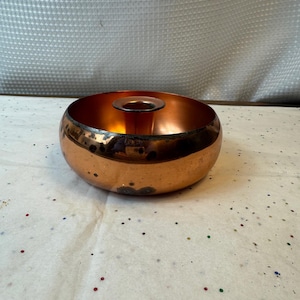 May include: A copper candle holder with a circular design. The candle holder has a wide, rounded base and a small, central opening for a candle. The copper has a warm, reddish-brown hue with some darker areas, giving it a vintage look.