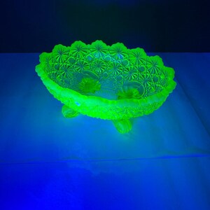 May include: A glowing green glass bowl with a detailed starburst pattern and three small feet. The bowl has a scalloped rim and is illuminated with a bright green light, creating a striking visual effect. The bowl is likely vintage.