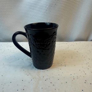 May include: A tall, black ceramic mug with a textured surface and a curved handle. The mug has a glossy finish and is designed for hot beverages. The mug is empty and sits on a white surface.