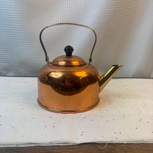Puede incluir: Una tetera de color cobre con asa y boquilla de latón. La tetera tiene un pomo negro en la tapa y un cuerpo redondeado. La boquilla está en ángulo y el asa se arquea sobre la parte superior.