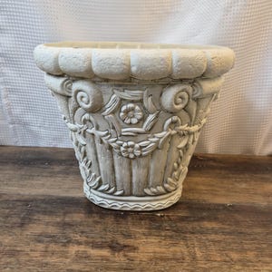 Puede incluir: Jardinera decorativa de color blanquecino con detalles florales y de volutas ornamentadas. La jardinera tiene un borde texturizado y un cuerpo estriado. El diseño incluye una flor central y guirnaldas. Fabricada con material duradero.