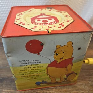 Könnte beinhalten: Vintage Winnie the Pooh Spieluhr. Die rot-weiße Box zeigt einen lächelnden Winnie the Pooh mit einer Trommel und einem roten Ballon. Die Oberseite hat ein Karneval-Design. Der Text auf der Seite lautet "But most of all Winnie the Pooh!"