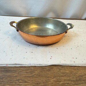 Puede incluir: Una sartén poco profunda de color cobre con dos asas. La sartén tiene forma redondeada y un interior brillante. Las asas están unidas a los lados de la sartén. La sartén está sobre una superficie blanca.