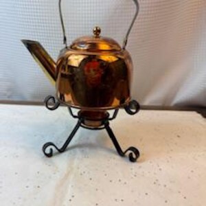 May include: A small, copper-colored tea kettle with a curved spout and a black metal stand. The kettle has a handle and a lid with a small knob. The stand has a decorative, scrolled design. The kettle is sitting on a white surface.