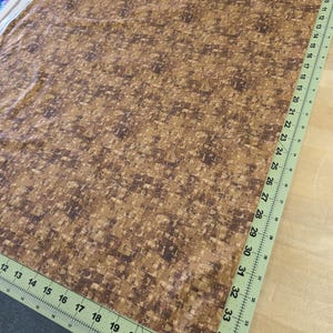 May include: Brown fabric with a repeating pattern of small, irregular rectangles in shades of brown and tan. A green ruler with inch measurements is along the edge of the fabric. The fabric is likely used for sewing or crafting projects.