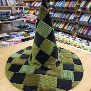 May include: A pointed hat constructed from a patchwork of square fabric pieces in shades of black, olive green, and lime green. The hat features a wide, circular brim and a tall, conical crown, with visible stitching outlining each square patch.