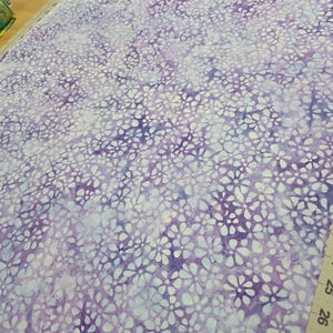 May include: A roll of fabric with a floral pattern in shades of purple and white. The fabric is spread out on a cutting board with a ruler showing measurements in inches. The floral design is a repeating pattern of small, stylized flowers.