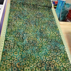 May include: Green fabric with a repeating pattern of gold and teal starburst shapes. The fabric is laid out on a wooden surface with a ruler along the edge. The fabric is likely used for quilting or sewing projects.