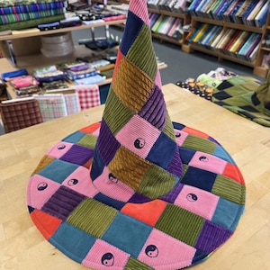 May include: A conical hat constructed from a patchwork of corduroy fabric squares in various colors including pink, blue, green, orange, and purple. The hat features a repeating black and white yin-yang symbol printed on several of the fabric squares.