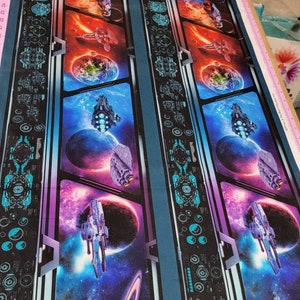May include: Fabric with a space-themed design. The fabric features a repeating pattern of spaceships, planets, and galaxies in vibrant colors. The design includes a dark blue border with a futuristic pattern.