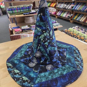 May include: A conical hat constructed from quilted fabric panels in shades of blue, teal, and black. The fabric features celestial patterns including crescent moons, stars, and abstract web-like designs, with visible stitching throughout the surface.