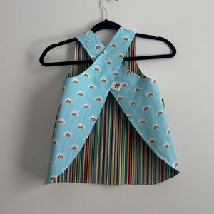 May include: A reversible apron with a blue background and rainbow pattern on one side and vertical rainbow stripes on the other. The apron is hanging on a black hanger.
