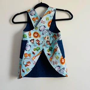 May include: A blue and white animal print apron with a cross-back design. The apron features a light blue background with various cartoon animals, including giraffes, lions, and elephants. Dark blue pockets and trim complete the look.