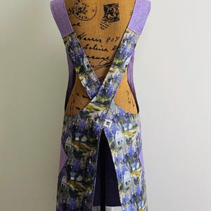 May include: A purple and white apron with a floral pattern featuring white houses and purple flowers. The apron has a criss-cross back strap and two pockets on the sides.