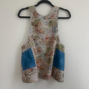 May include: Apron featuring a Winnie the Pooh print with blue pockets. The apron is made of a light-colored fabric with a repeating pattern of Winnie the Pooh characters in a forest setting. The pockets are a solid blue color.