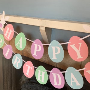 May include: A pastel-colored Happy Birthday banner made of paper eggs with white letters. The banner is strung together with white ribbon.
