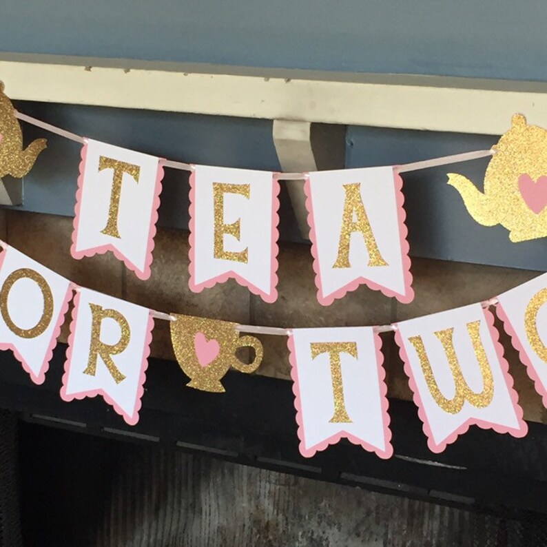 May include: A pink and gold banner with the words "Tea for Two" in gold glitter letters. The banner has a scalloped edge and is decorated with pink and gold teapots and teacups.