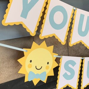 May include: A banner with the words "YOU SUNSHINE" in light blue letters on white pennants with yellow scalloped edges. A smiling yellow sun with a blue bow tie is also featured.
