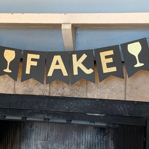 May include: Black and gold banner with the word "FAKE" and wine glass silhouettes. The banner is hanging from a fireplace mantel.