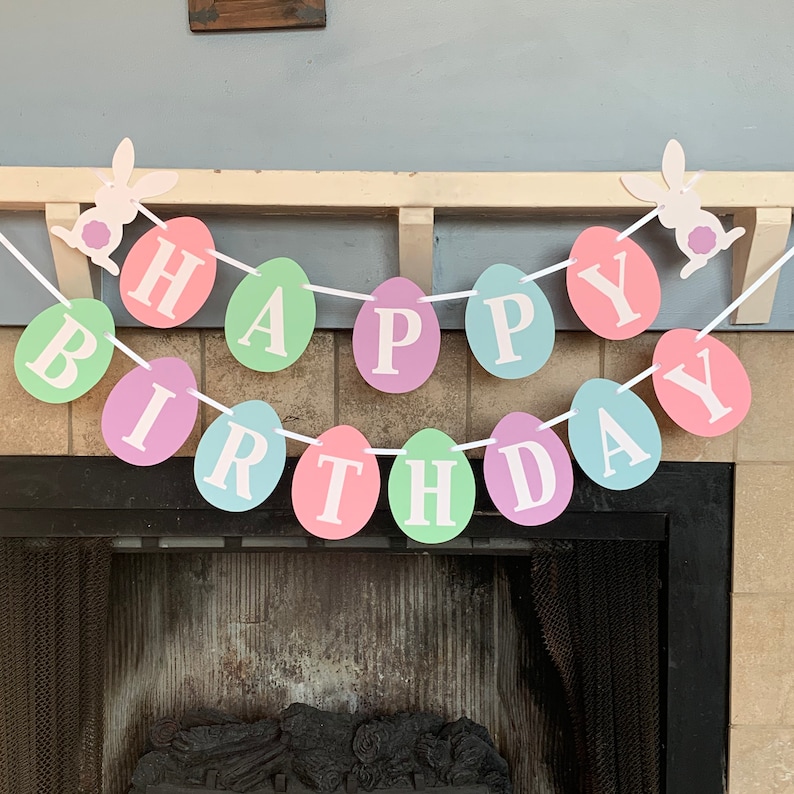May include: A pastel colored Happy Birthday banner with white letters and bunny rabbit cutouts. The banner is made of paper and is strung together with white ribbon.