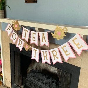 May include: A pink and gold banner with the words "Tea for Three" in gold glitter letters. The banner is decorated with pink and gold teapots and hearts.